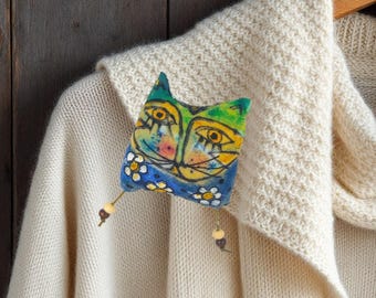 May include: A cream-colored knit cardigan featuring a brooch shaped like a stylized cat head. The cat is painted in yellow, green, and blue with white flower accents and has two dangling wooden beads attached to the bottom corners of the piece.