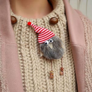 May include: A whimsical brooch featuring a fluffy, gray bird with googly eyes and a red and white striped hat. The bird is attached to a beige sweater and a pink coat. The brooch has dangling beads.