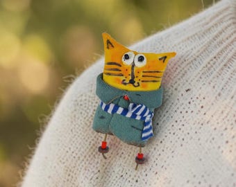 May include: A handmade cat brooch with an orange head, black stripes, and a green jacket. The cat wears a blue and white striped scarf and has small dangling legs with red and brown beads. The brooch is pinned to a cream-colored sweater.