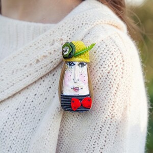 May include: A whimsical handmade brooch featuring a painted face with a yellow hat, green accents, and a red bow tie. The brooch has long, light brown hair and a striped shirt. This unique accessory adds a touch of artistic flair.