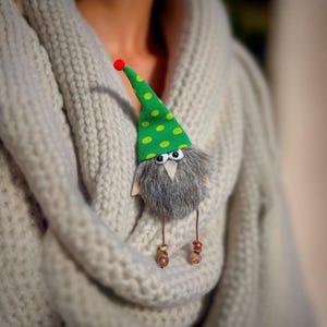 Puede incluir: Broche artesanal caprichoso con forma de gnomo. Tiene un gorro verde con lunares amarillos, un cuerpo peludo gris y patas de madera con cuentas en los pies. El broche está prendido a una bufanda de punto gris claro.