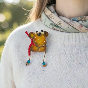 Könnte beinhalten: Eine handgefertigte Brosche in Form eines gelb-braunen Tieres mit weißen Punkten. Die Brosche hat ein rotes Band und hängende Beine mit blauen Perlen. Sie ist an einem weißen Pullover befestigt, im Hintergrund ist ein floraler Schal zu sehen.