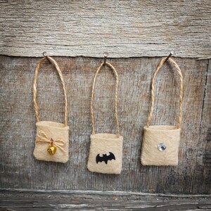 Op de afbeelding: Drie kleine jute zakjes hangen aan spijkers aan een houten muur. De zakjes zijn versierd met een bel, een vleermuis en een strass steentje.