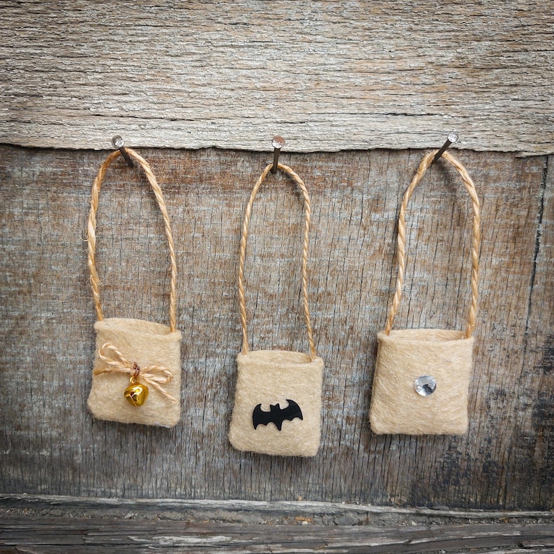 Op de afbeelding: Drie kleine jute zakjes hangen aan spijkers aan een houten muur. De zakjes zijn versierd met een bel, een vleermuis en een strass steentje.
