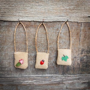 Op de afbeelding: Drie kleine jute zakjes hangen aan spijkers aan een houten muur. Elk zakje heeft een ander ontwerp: een rode lieveheersbeestje, een roze bloem en een groen blad.