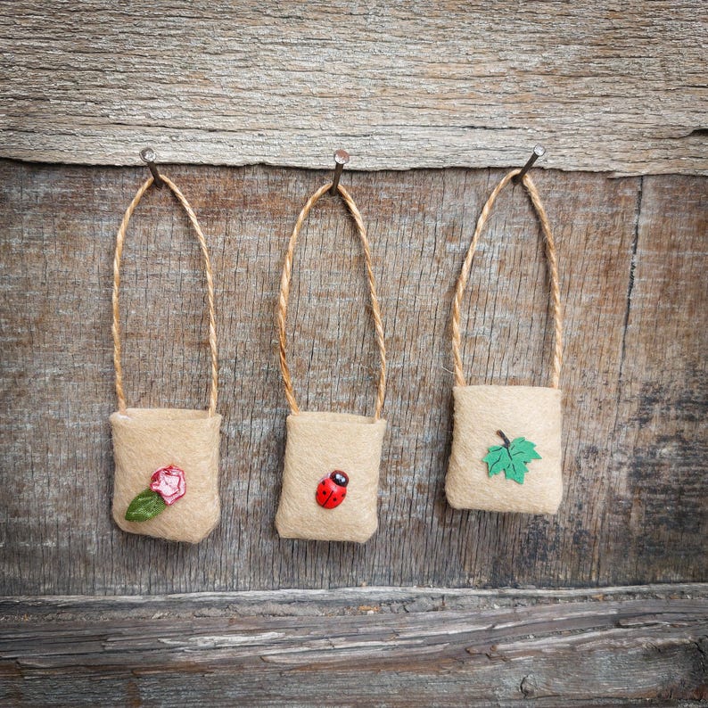Op de afbeelding: Drie kleine jute zakjes hangen aan spijkers aan een houten muur. Elk zakje heeft een ander ontwerp: een rode lieveheersbeestje, een roze bloem en een groen blad.