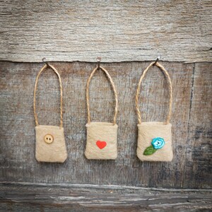 Op de afbeelding: Drie kleine jute zakjes hangen aan spijkers aan een houten muur. De zakjes zijn versierd met een rood hart, een blauwe bloem en een bruine knoop.