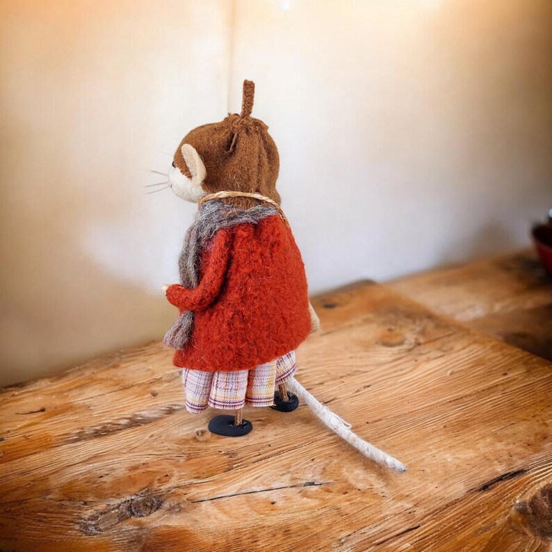 Op de afbeelding: Een bruine en witte viltmuis met een rode jas, een bruine hoed en een geruite rok. De muis heeft een lange witte staart.