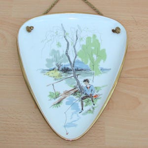 Może przedstawiać: Vintage, trójkątna płyta ścienna z namalowaną sceną osoby łowiącej ryby. Biały ceramiczny element ma złote obramowanie i pleciony sznur do zawieszenia. Grafika przedstawia drzewo, wodę i postać w kolorze niebieskim. Wysokość: około 20 cm.