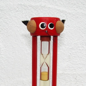 Peut inclure: Un sablier rouge avec un visage de chat sur le dessus. Le sablier a des oreilles noires, des yeux blancs et un nez noir. Le sable est jaune et le verre est transparent.