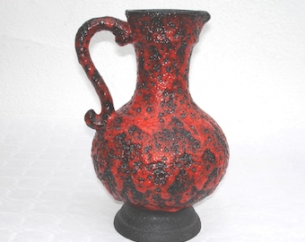 May include: Red and black ceramic pitcher with a textured surface. The pitcher has a wide base and a curved handle.
