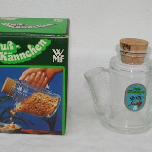 Peut inclure: Un récipient en verre transparent en forme de théière avec un bouchon en liège, à côté d'une boîte verte. La boîte présente le texte "Nuß Kännchen" et une illustration du récipient. Le récipient a un autocollant bleu et blanc.