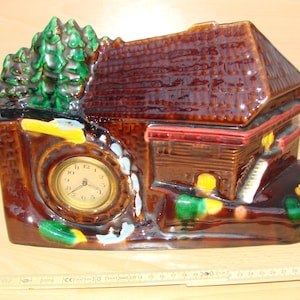 May include: A ceramic clock shaped like a cabin with a water wheel and trees. The clock face is gold with black numbers and hands. The cabin is brown with a red roof and green and yellow accents.