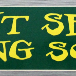 May include: A rectangular sign with a green background and yellow text that reads "PLANT SEEDS & SING SONGS". The sign has a white border and a gray wooden frame.