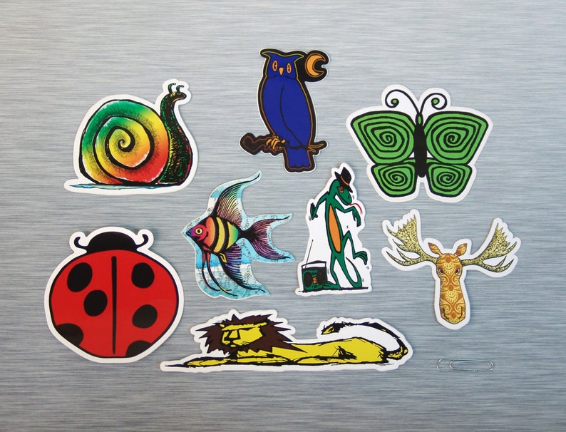 May include: A collection of colorful stickers featuring various animals and insects. Includes a snail with a rainbow shell, a ladybug, a blue owl, a green butterfly, a fish, a lizard, a moose head, and a lion. The stickers are on a silver surface.