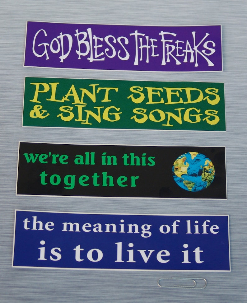 May include: Four colorful stickers with different messages. The top sticker is purple with white text that reads "God Bless The Freaks". The second sticker is green with yellow text that reads "Plant Seeds & Sing Songs". The third sticker is black with a blue and green globe image and white text that reads "we're all in this together". The bottom sticker is blue with white text that reads "the meaning of life is to live it".