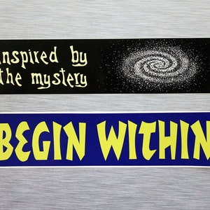 May include: Two stickers with inspirational messages. The top sticker is black with a white galaxy image and the text "inspired by the mystery". The bottom sticker is blue with yellow text that reads "BEGIN WITHIN".