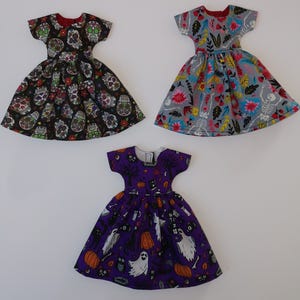 May include: Three doll dresses with different Halloween-themed prints. The first dress is black with a skull and flower pattern. The second dress is gray with a floral and skeleton pattern. The third dress is purple with a ghost, pumpkin, and cat pattern.