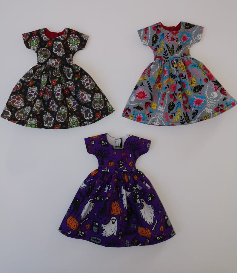 May include: Three doll dresses with different Halloween-themed prints. The first dress is black with a skull and flower pattern. The second dress is gray with a floral and skeleton pattern. The third dress is purple with a ghost, pumpkin, and cat pattern.