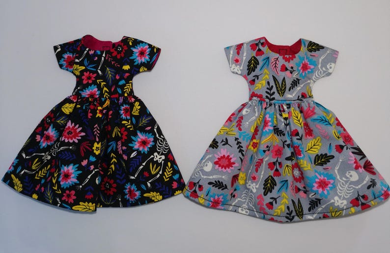 May include: Two doll dresses with a floral and skeleton print. One dress is black with pink, yellow, and blue flowers. The other dress is gray with pink, yellow, and blue flowers.