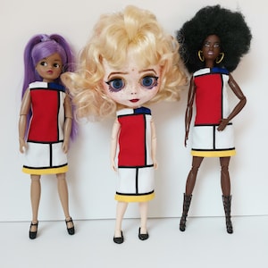 Op de afbeelding: Drie modepoppen gekleed in Mondriaan-stijl jurken. De jurken hebben een rood, wit en zwart kleurblokontwerp met een gele zoom. De poppen hebben verschillende huidtinten en kapsels, waaronder paars haar, blonde krullen en een zwarte afro.