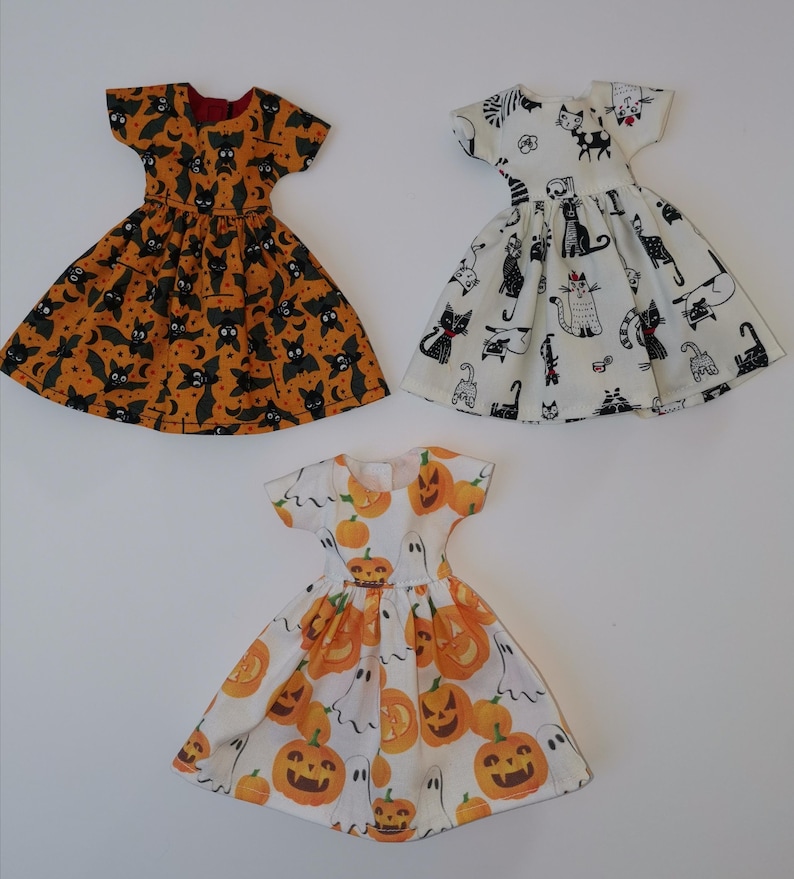 May include: Three Halloween-themed doll dresses. One dress is orange with black bats, one is white with black cats, and one is white with orange pumpkins and ghosts.