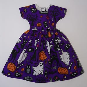 May include: A purple dress with a Halloween print featuring black cats, ghosts, pumpkins, and other spooky elements.