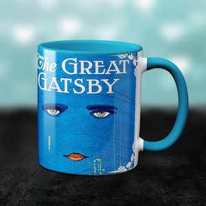 May include: A blue and white ceramic mug with a teal rim and handle. The mug features a vintage illustration of a woman's face with the text "The Great Gatsby" printed on the front.