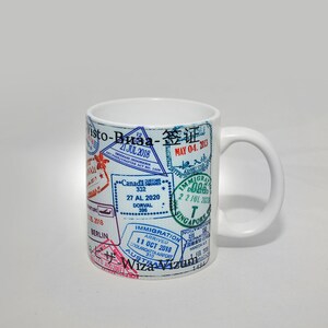 May include: White ceramic mug with a design of various colorful passport stamps, including "Visa", "签证", "May 04, 2013", "21 Jul 2018", "Canada", "27 AL 2020", "Dorval", "Immigration", "11 Oct 2018", "Coolangatta Airport", "Australia", "Wiza", "Singapore", "22 Jul 2020", and "Haneda Airport".