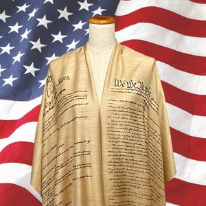 Puede incluir: Un chal beige con el texto de la Constitución de los Estados Unidos impreso en tinta negra. El chal está drapeado sobre un maniquí con un fondo de bandera estadounidense.