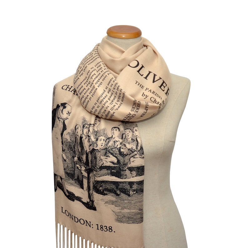 May include: A beige scarf with a black and white illustration of a scene from the novel Oliver Twist by Charles Dickens. The text "Oliver Twist" and "The Parish" are printed on the scarf. The text "London: 1838" is printed at the bottom of the scarf.