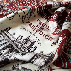 May include: A cream silk scarf featuring the text "Pride and Prejudice, Pemberley" alongside detailed illustrations in burgundy. The design includes architectural drawings and floral patterns. The scarf is draped, showing its flowing texture and intricate design.