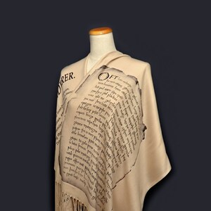 May include: A beige shawl with a fringe border. The shawl features a printed design of black text in an old-fashioned font. The text reads "OFT..."