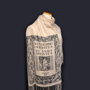 May include: A cream-colored scarf with a black and white print of the book cover for "Wuthering Heights" by Emily Bront&euml;. The print includes the title, author, and publication date of 1847. The scarf has fringe on the bottom.