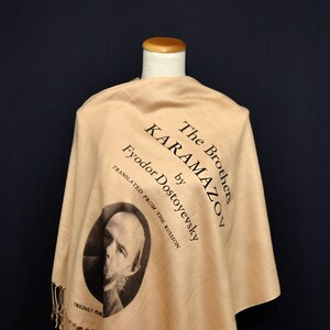 May include: A beige shawl with a black print of the book "The Brothers Karamazov" by Fyodor Dostoevsky. The print includes a portrait of the author and the text "Translated from the Russian" and "Originally Published".