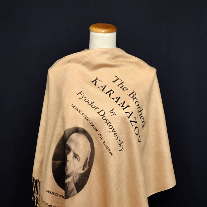 May include: A beige shawl with a black print of the book "The Brothers Karamazov" by Fyodor Dostoevsky. The print includes a portrait of the author and the text "Translated from the Russian" and "Originally Published".