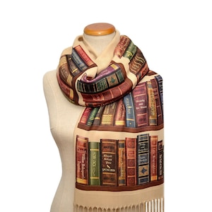 Op de afbeelding: Een beige sjaal met een boekenkastprint met de titels van klassieke boeken, waaronder "Pride and Prejudice", "Moby Dick", "Peter Pan", "The Call of the Wild" en "Les Misérables".