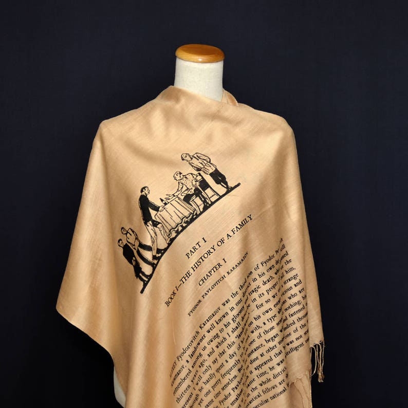 May include: A beige silk scarf with a black and white illustration of people sitting at a table. The scarf has text printed on it that reads "Part I Book I - The History of a Family Chapter 1 Fyodor Pavlovich Karamazov was the..."