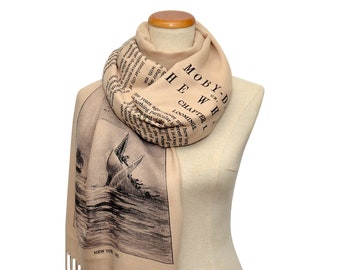 Op de afbeelding: Een beige sjaal met een zwart-witte print van een walvis en tekst uit het boek Moby Dick. De tekst luidt "MOBY-DICK or THE WHALE CHAPTER L LOOMINGS". De sjaal heeft franjes aan de onderkant.