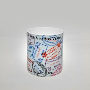 May include: White ceramic mug with a design of various passport stamps from different countries, including Paris, Hawaii, Tokyo, and Berlin. The stamps are in blue, red, and black.