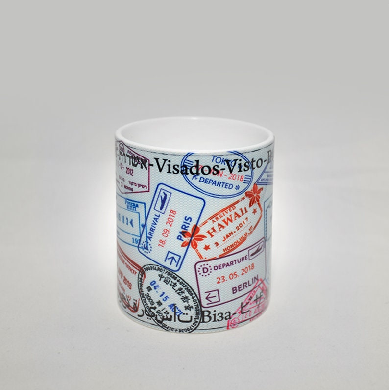May include: White ceramic mug with a design of various passport stamps from different countries, including Paris, Hawaii, Tokyo, and Berlin. The stamps are in blue, red, and black.
