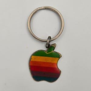 Peut inclure: Un porte-clés vintage avec un motif de pomme aux couleurs de l'arc-en-ciel. La pomme est rayée de vert, jaune, orange, rouge et bleu. Le porte-clés a un anneau argenté et un petit anneau de connexion.