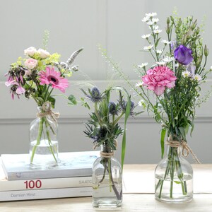 May include: Three glass vases containing different types of flowers. The vase on the left holds pink and white flowers, the middle vase holds blue and purple flowers, and the vase on the right holds pink and white flowers.