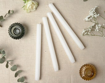 May include: Four white taper candles arranged in a fan shape on a beige linen surface. Two glass candle holders are also visible, one green and one brown.