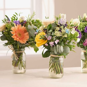 May include: Three glass jars filled with colourful bouquets of flowers. The jars are tied with twine and the flowers include pink, yellow, orange, white, purple, and blue blooms.