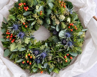 May include: A circular wreath composed of various green foliage including pine needles, eucalyptus leaves, and oak leaves. The arrangement is accented with bright red berries and spiky, vibrant blue thistle flowers distributed throughout the greenery.