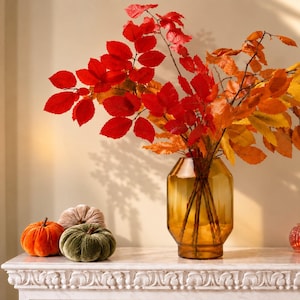 May include: A tall, amber-coloured glass vase with a faceted, hexagonal body holds a large arrangement of vibrant autumn branches. The branches feature bright red and orange leaves. Three small, plush fabric pumpkins in orange, olive green, and beige sit nearby.