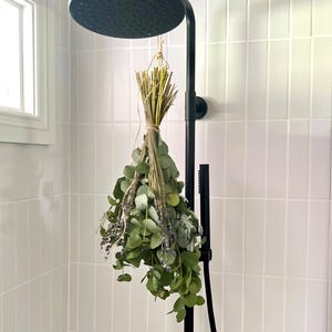 May include: A bunch of fresh eucalyptus and lavender suspended from a black shower head. The leaves are green and the lavender is purple. The shower fittings are black, and the walls are tiled white.