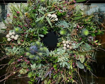 May include: A vibrant, circular wreath composed of various green and blue-green foliage, including thistles, eucalyptus, and sprigs of berries. The wreath is adorned with small, round, off-white accents and delicate brown twigs, creating a natural, textured appearance.
