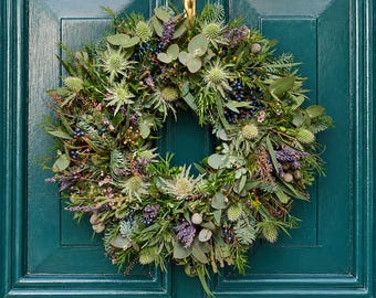 May include: A circular wreath composed of green eucalyptus leaves, spiky green sea holly, purple lavender sprigs, and dark blue berries. The foliage is arranged in a dense, textured pattern, featuring various shades of green and purple accents.